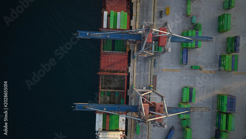 Mercante en muelle de carga junto a grúa y camines cargando contenedores en el barco , vista aérea cenital