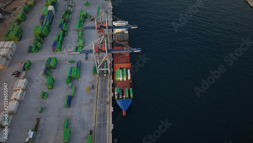 Mercante en muelle de carga junto a grúa y camines cargando contenedores en el barco , vista aérea cenital
