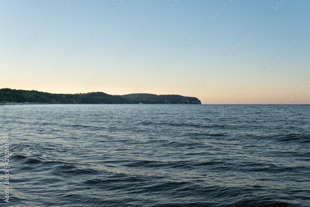 Obraz premium Tranquil Ocean Horizon: A vast expanse of deep blue ocean meets a clear, pale sky, with a distant, tree-lined coastline fading into the horizon.