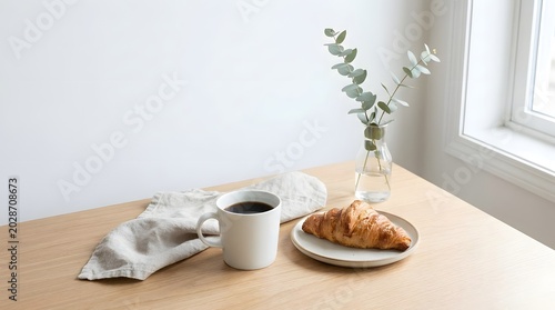 A minimalist breakfast scene with coffee, a croissant, and eucalyptus on a light wooden table.
