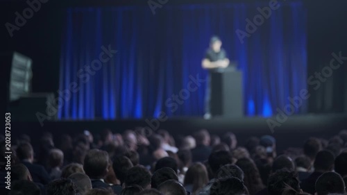 Large audience watching lone speaker deliver presentation on dimly lit stage, corporate conference atmosphere, focus on crowd and event.
