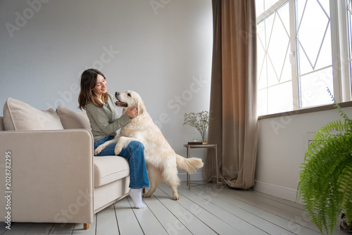 Billede på lærred Young woman sitting on sofa petting golden retriever standing on hind legs with paws up, warm moment in bright cozy living room