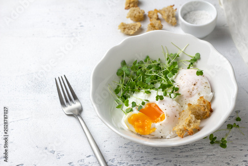 Poached eggs with microgreens and bread croutons in white bowl on light rustic background