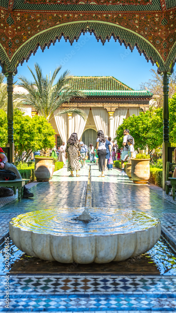 Fototapeta premium People walk through the Secret Garden in Marrakesh, Morocco. Sunlight shines down on the garden as visitors enjoy the lush plants and scenic views of architecture.