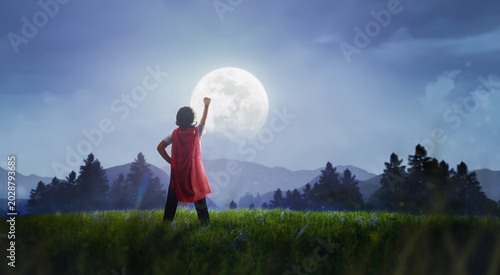 Young boy in red superhero cape raising fist towards a giant full moon at night