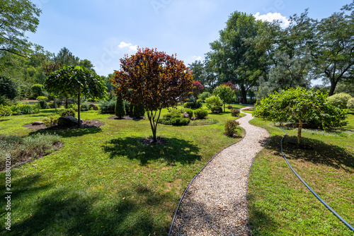 A winding gravel pathway curves through a landscaped public park with manicured green lawns, young ornamental trees with green and reddish foliage, and mature trees under a clear blue sky