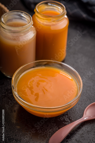 Creamy Pumpkin Soup In Glass Bowl With Jars Of Puree And Spoon On Dark Surface