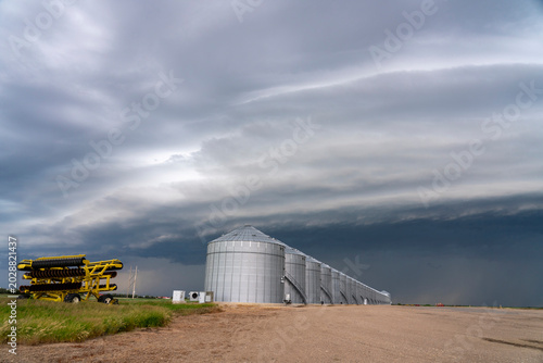 Agricultural grain silos stand under a dramatic, stormy sky with a farm implement nearby