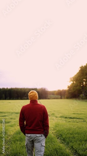 Cinematic footage of a man standing alone in a wide green field during golden hour while facing the horizon and surrounding nature. Peaceful countryside scenery conveys freedom, reflection.