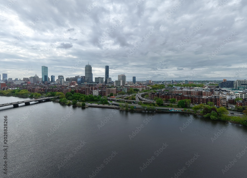 Obraz premium Aerial drone view of the Boston skyline under a cloudy sky, featuring the Charles River, Harvard Bridge, and Back Bay brownstones leading toward the Prudential and John Hancock towers.