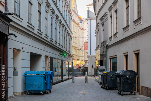 Fotografi Empty European Alley With Trash Bins and Pharmacy Green Cross Sign on Narrow Cit