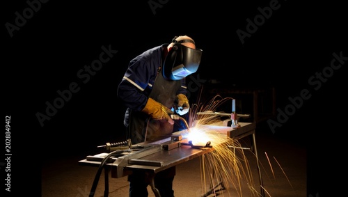 Fotografie A welder in a dark auto-darkening helmet and blue work jacket leans over a steel plate on a metal workbench