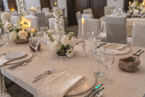 Elegant wedding reception table setting with white floral centerpieces, candles and stone chargers. Luxury banquet dinner decor at wedding venue.
