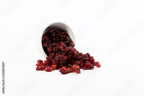 Dried cranberries spilled out from a tipped white ceramic cup onto a clean white background