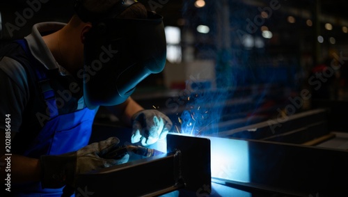 Bild auf Leinwand Striking chiaroscuro composition of a young welder in a blue apron bending close over a steel beam corner, the vivid blue arc light cutting through darkness with blue smoke and sparks rising softly
