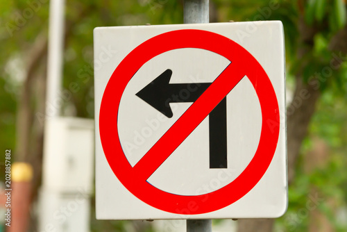 No left turn sign on white traffic sign with red circle and diagonal slash, outdoor road safety symbol with blurred green background