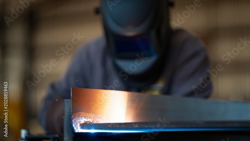 Tableau sur toile Shallow focus shot of a freshly formed weld bead glowing orange on a steel girder edge, with the helmeted welder in gray worksuit softly blurred in the warm background of an industrial workshop