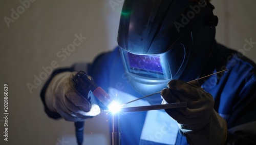 Konstfotografi Close portrait of a welder in blue work jacket and large auto-darkening helmet with glowing purple visor reflection, TIG welding with a filler rod with bright white arc in a dim workshop