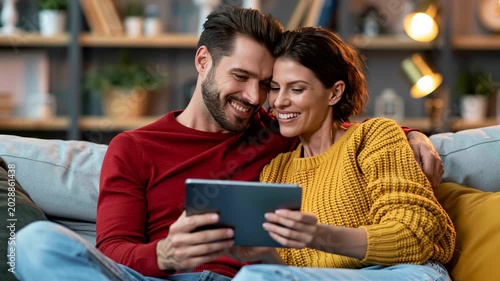 This depicts a happy couple enjoying leisure time and digital connection at home. A smiling man and woman are relaxing comfortably on a sofa, sharing a tablet for online entertainment. This scene conv