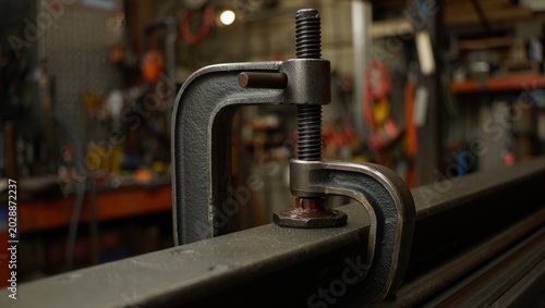 Foto Macro close-up of a heavy cast iron G-clamp secured onto a steel rail in a workshop, textured metal surface and threaded screw in sharp focus with colorful blurred workshop shelving behind