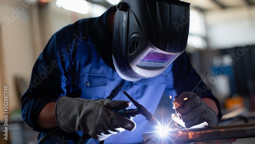 Foto Close-up of a professional TIG welder in blue work shirt and large black auto-darkening helmet with gray TIG gloves welding a precise metal joint with a bright white arc in a workshop