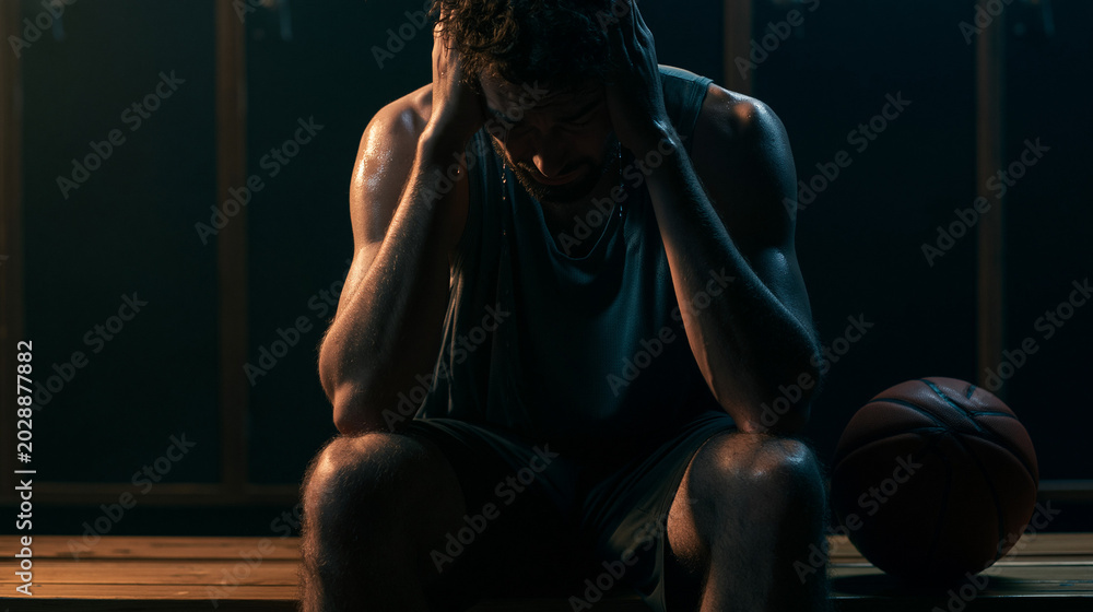 Fototapeta premium Devastated male basketball player sitting in a dark locker room crying. Concept of sports failure, getting fired from the team, and mental exhaustion