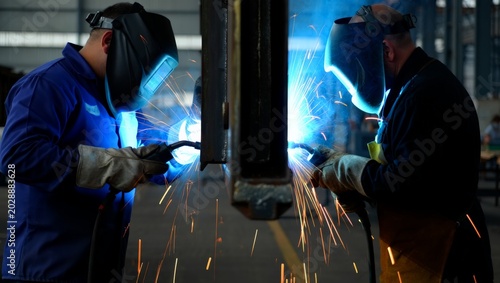Tableau sur toile Two professional welders in matching blue uniforms and auto-darkening helmets working simultaneously on opposite sides of a steel girder with vivid blue arcs and orange sparks in a factory