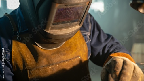 Quadro em tela Atmospheric portrait of a welder wearing a large auto-darkening helmet and brown leather welding apron over a blue uniform, leaning forward in warm hazy workshop light with soft background bokeh