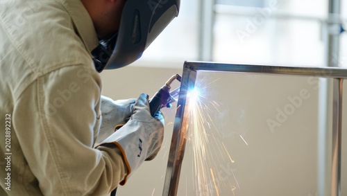 Schilderij op canvas Professional welder in a cream work coverall and dark welding helmet welds a steel frame corner with a MIG torch, bright blue arc and golden sparks flying in a clean studio workshop