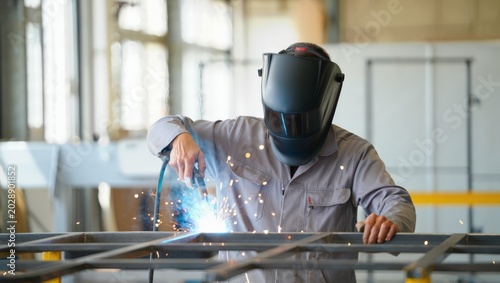 Quadro em tela Front-facing studio portrait of a welder in grey coverall and dark helmet MIG welding a steel grid frame with a bright blue arc and sparks in a naturally sunlit industrial workshop environment