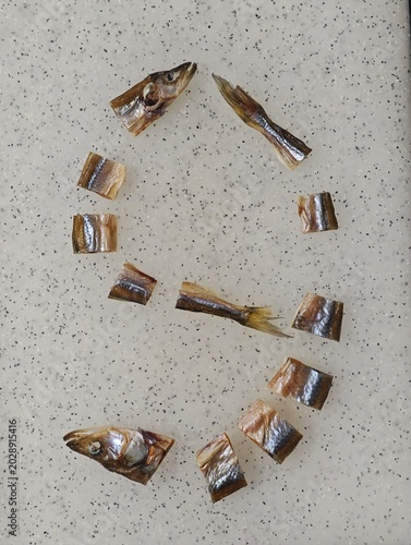 The number 9, a Ukrainian Cyrillic digit, is creatively formed on a speckled light gray surface using an assortment of dried smelt fish pieces, including its distinct head and tail, arranged in an