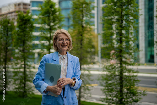 Konstfotografi Mature businesswoman holding laptop walking in city park
