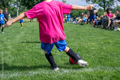 Youth soccer player kicks ball on the field during afternoon game with spectators in the background watching closely