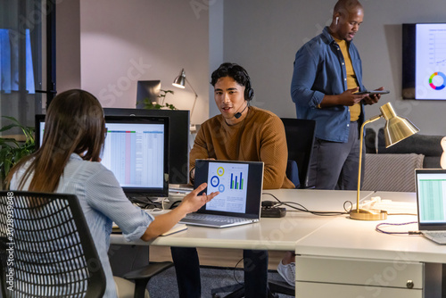 Diverse coworkers collaborating around shared desks in open-plan office with laptops headset tablet