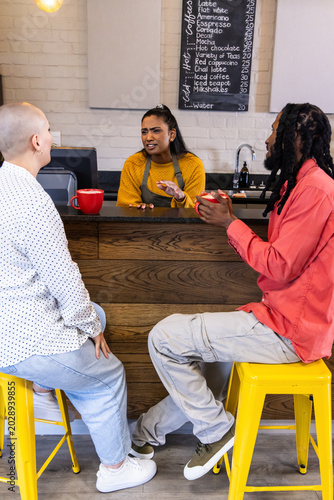 Indian barista in mustard top speaking, gesturing behind cafe bar while customers holding red mugs