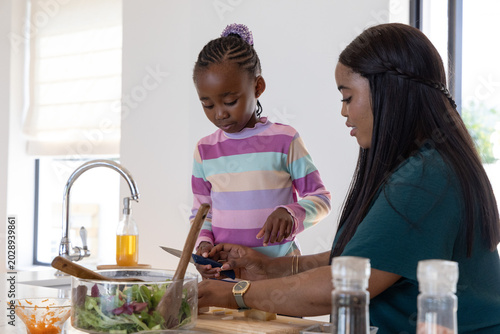 African American mother and daughter preparing salad at kitchen countertop with glass bowl, peeler