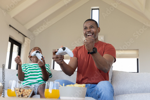 Foto African American father and son cheering, holding white controllers on sofa with