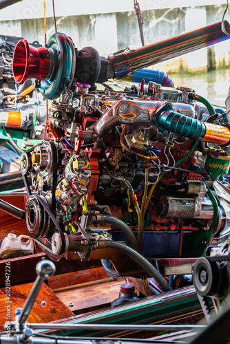 A high performance turbocharged engine sits prominently on a traditional Thai longtail boat. The mechanical assembly features vibrant metallic colors and intricate custom components in a close up view
