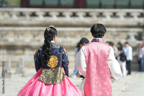Two people in traditional Korean attire walk away from the camera. Their backs show ornate patterns and bright colors. Woman wears a pink skirt with gold embroidery