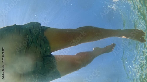Underwater shot looking up at a man in swim trunks casually swimming by blowing bubbles on a sunny day with blue sky. 