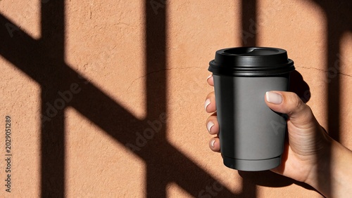 Hand holding grey paper coffee cup with black lid mockup against terracotta wall with dramatic window shadows