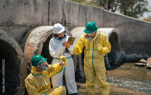 Scientists in protective hazmat suits analyzing contaminated runoff water from concrete sewer outlets during environmental inspection and industrial pollution control operation outdoors.