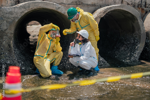 Scientific inspection team analyzing hazardous wastewater collected from polluted coastal drainage canal while monitoring ecological damage and industrial contamination sources nearby.