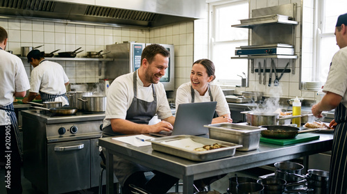 Restaurant Kitchen Collaboration: Two chefs are working together on a laptop in a busy professional kitchen, surrounded by culinary activity, conveying teamwork and modern kitchen management.