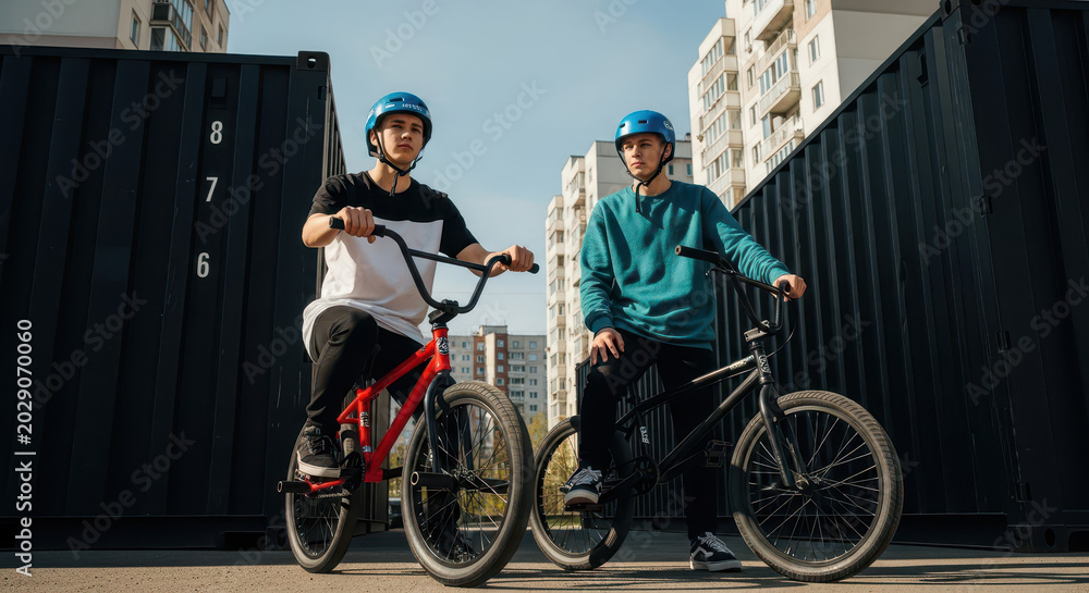 Fototapeta premium Two young men posing with BMX bikes in an urban industrial setting with shipping containers and city buildings.