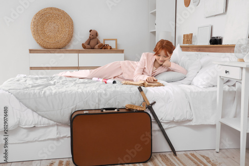 Cute teenage happy redhead girl in pajamas with world map reading adventure b...