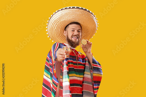 Konstfotografi Handsome young man in Mexican poncho and sombrero holding shot of tequila on yel