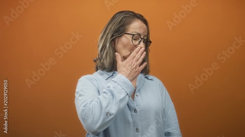 Woman grey hair senior covers mouth with hand in a studio with orange backdrop, wearing glasses, light blue shirt and gold necklace; disgust discomfort.