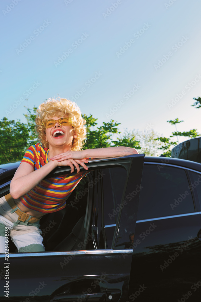 Fototapeta premium Joyful woman leans from a car window, laughing with a retro hairstyle and striped shirt, bright outdoor scene, cinematic color, casual lifestyle moment, playful mood.