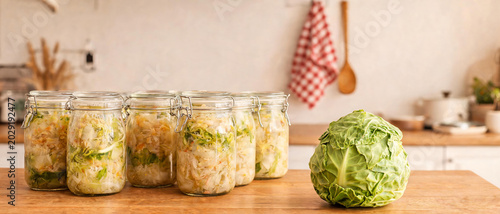 Obraz w ramie Row of glass jars with homemade sauerkraut and a fresh head of cabbage on a wooden table in a light kitchen.